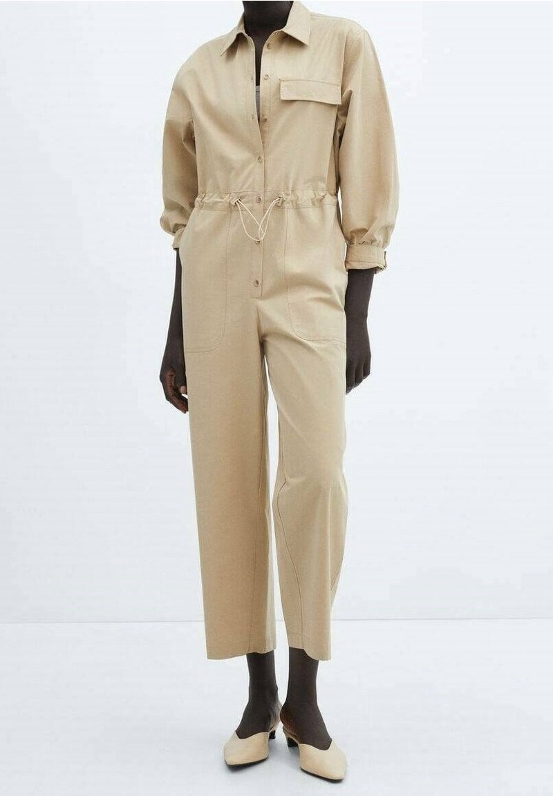 Mannequin portant une combinaison beige à manches longues avec taille à cordon, poche poitrine, et chaussures beiges à petit talon sur fond blanc.