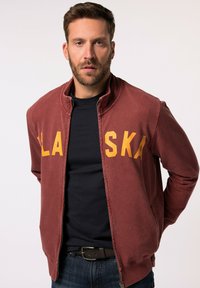 Giacca zip-up color bordeaux con scritta "ALASKA" gialla, tessuto morbido, colletto a coste e maniche lunghe. Indossata sopra una maglietta nera.