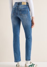 Jean bleu slim avec poches arrière porté par une personne portant un haut blanc à manches longues et des baskets blanches avec des accents verts.