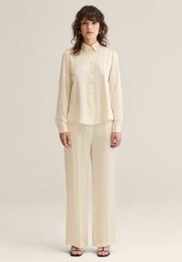Kvinde med krøllet hår iført cremefarvet skjorte med knapper og matchende bukser med vide ben, stående foran en ensfarvet baggrund.