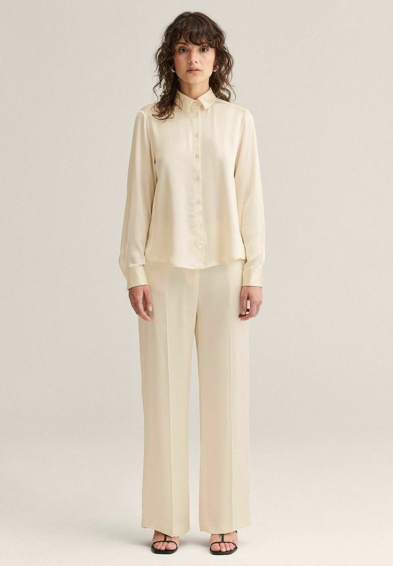 Kvinde med krøllet hår iført cremefarvet skjorte med knapper og matchende bukser med vide ben, stående foran en ensfarvet baggrund.