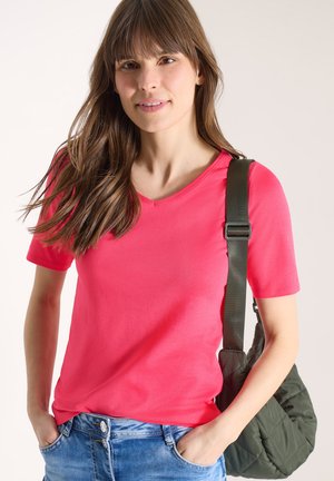 Femme aux longs cheveux bruns portant un T-shirt corail à col en V et un jean bleu, avec un sac à bandoulière matelassé vert foncé.
