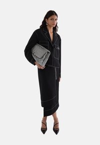 Zwarte getailleerde blazer met witte stiksels, gecombineerd met een bijpassende rok. Houdt een gequilt grijze handtas vast en draagt zwarte puntige hakken.