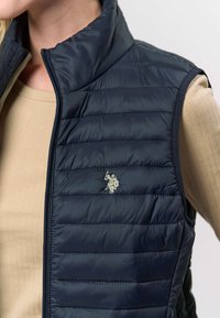 Mornarsko modra puhasta telovnik z visokim ovratnikom, s horizontalno prešito teksturo in srebrnim vezenim logotipom na levi strani prsnega koša.