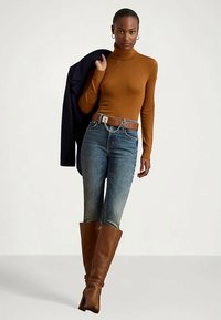 Brązowy sweter z golfem, niebieskie dżinsy z wysokim stanem, brązowy pasek z metalowymi akcentami, brązowe botki za kolano, granatowa kurtka zarzucona na ramię.