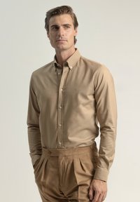 Camisa de botones color beige con cuello, de mangas largas y un diseño ajustado. Llevada con pantalones beige con pliegues a juego. Textura suave.