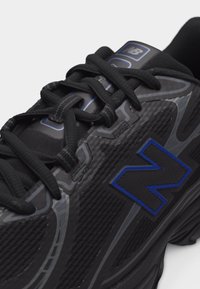 Μαύρο αθλητικό παπούτσι New Balance με λογότυπο "N" με μπλε περίγραμμα και μαύρα κορδόνια, που δείχνει υφασμένη λεπτομέρεια στη πλευρά και στην επάνω όψη.