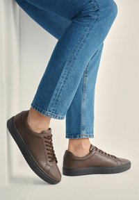 Osoba w niebieskich dżinsach i ciemnych brązowych skórzanych trampkach, jedna stopa oparta o białą ścianę na neutralnym tle.