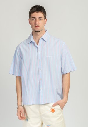 Man wearing light blue short-sleeve striped button-up shirt and off-white shorts with colorful embroidered designs, standing against plain background.