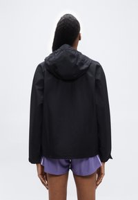 Femme aux longs cheveux portant une veste noire à capuche et un short de sport violet, debout de dos sur un fond clair uni.