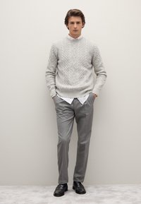 Grauer Kabelstrickpullover mit Rundhalsausschnitt, über einem weißen Hemd getragen, kombiniert mit grauen Hosen und schwarzen Slippern, vor einem neutralen Hintergrund stehend.