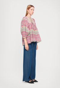 Blusa sovradimensionata multicolore con motivo floreale, maniche arricciate e scollo a chiave, abbinata a pantaloni in denim blu a gamba larga e scarpe nere.