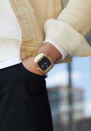 Goldener Metallarmbanduhr mit schwarzem quadratischen Ziffernblatt, rechteckigem Gehäuse und Gliederarmband. Am Handgelenk getragen, zeigt sie ein lässiges Outfit mit einer leichten Jacke.