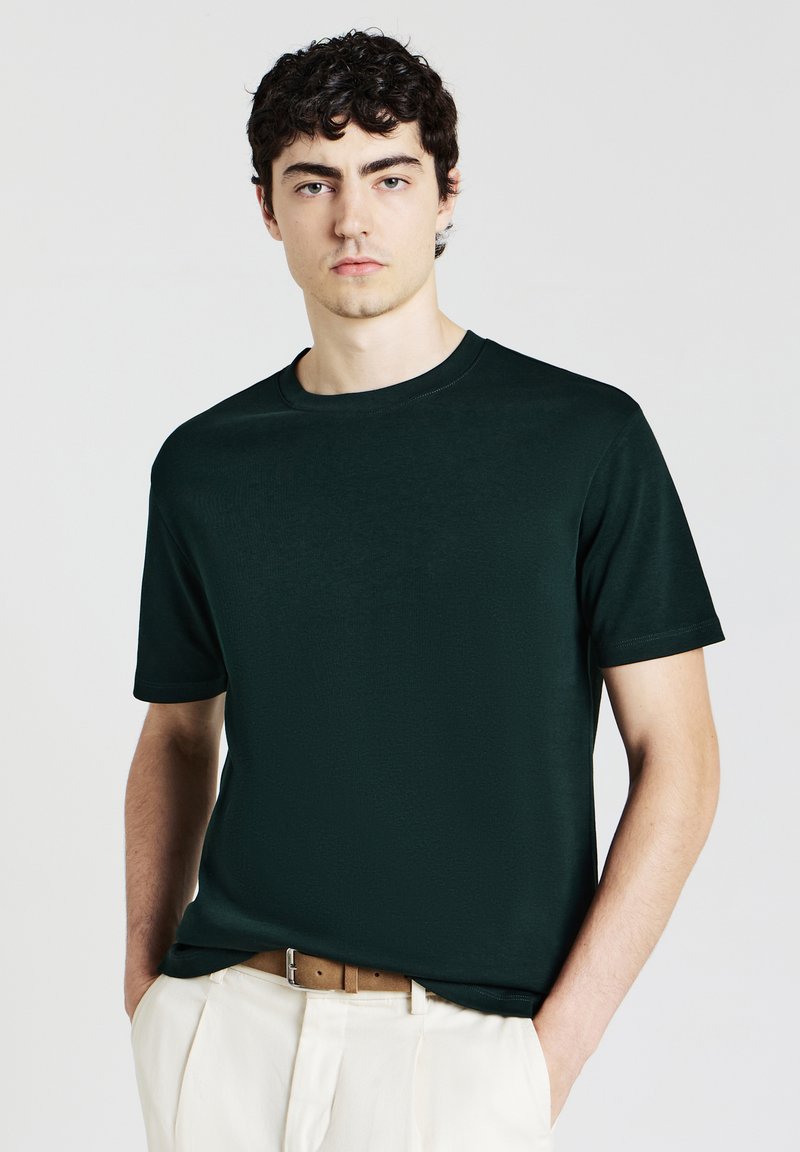 Giovane uomo che indossa una maglietta a maniche corte verde scuro infilata in dei pantaloni bianchi con una cintura marrone, in piedi con le mani nelle tasche.