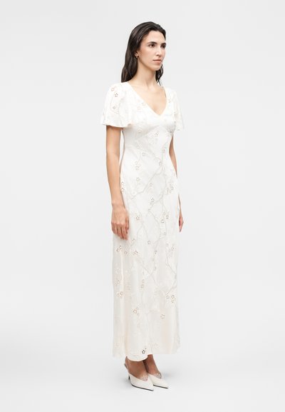 Γυναίκα που φοράει λευκό φόρεμα μέχρι τους αστραγάλους με κοντά μανίκια και κεντήματα με λουλουδένιες διακοσμήσεις, στέκεται μπροστά σε απλό φωτεινό φόντο.