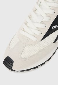 Lys grå semsket sneaker med hvite mesh-paneler, sorte detaljer og hvite snøringer. Har en teksturert såle og logo detalj nær hælen.