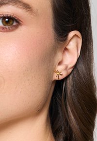 Złote kolczyki w kształcie małych kokardek, z gładką, błyszczącą powierzchnią i misternymi detalami na pętli i końcach.