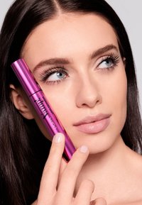 Tubetto di mascara cilindrico rosa tenuto vicino al volto di una modella, che mostra ciglia lunghe e scure e un colore naturale delle labbra. Si vede una pelle dalla texture liscia.
