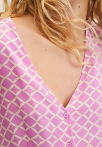 Blouse rose boutonnée avec un décolleté en V et un motif à grille blanche. Confectionnée dans un tissu lisse avec une texture douce.