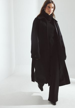 Čierny vonkajší kabát s veľkým golierom, dlhými rukávmi a bočnými viazaniami. Nosený cez čierny rolák a podobné priliehavé nohavice, doplnený o špičaté čierne topánky.