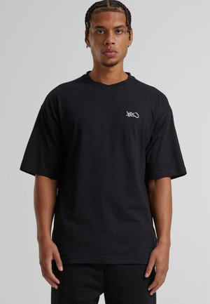 Black cotton t-shirt with short sleeves, oversized fit, featuring a small white logo on the left chest area. Smooth texture, simple design.