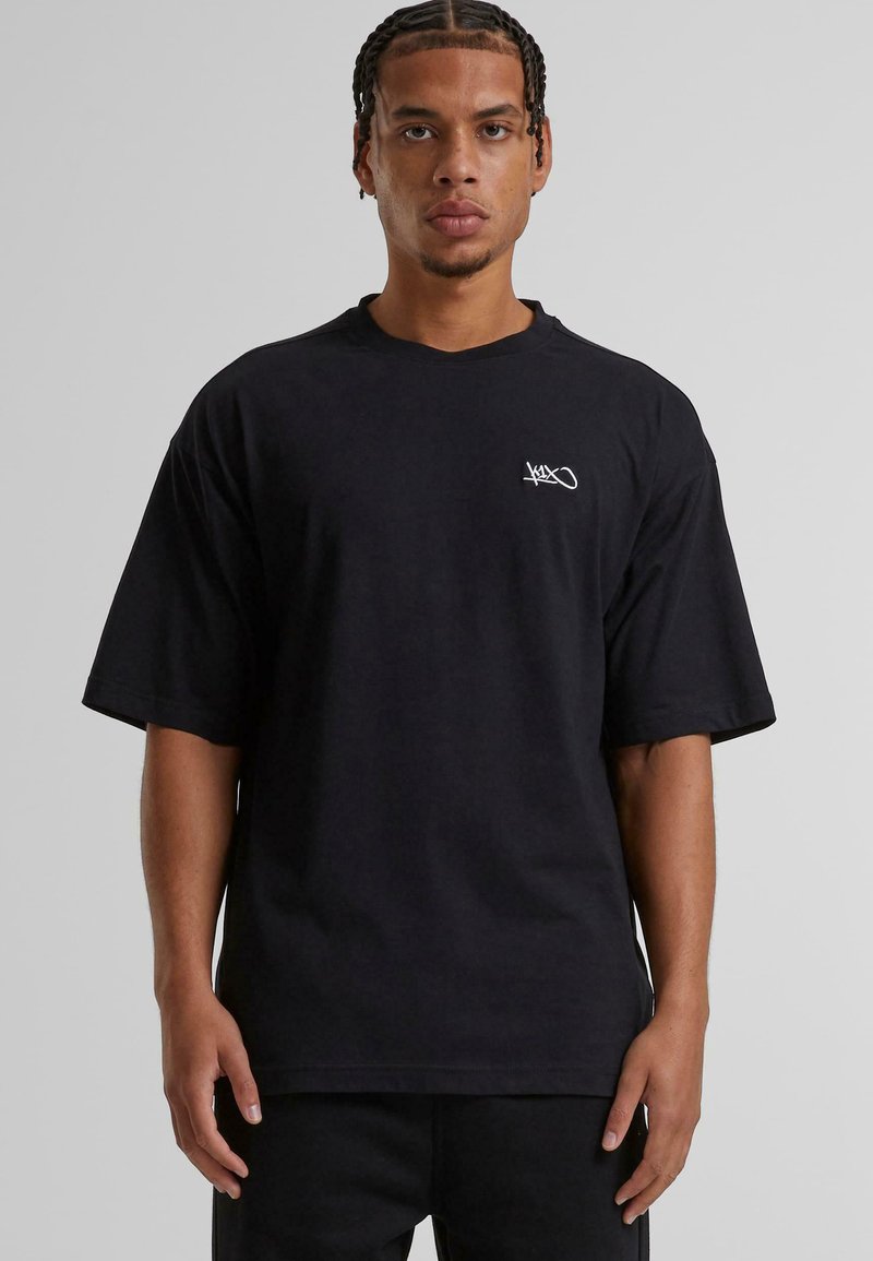 Camiseta de algodón negra con mangas cortas, corte oversized, que presenta un pequeño logo blanco en la parte izquierda del pecho. Textura suave, diseño sencillo.