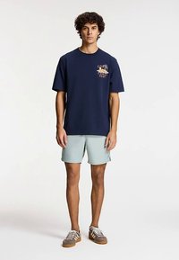 Tumesinine puuvillane lühikeste varrukatega t-särk, millel on rinnanäärmel värvikas logo. Kombineeritud heleroheliste lühikeste pükstega ja pruunide tossudega.