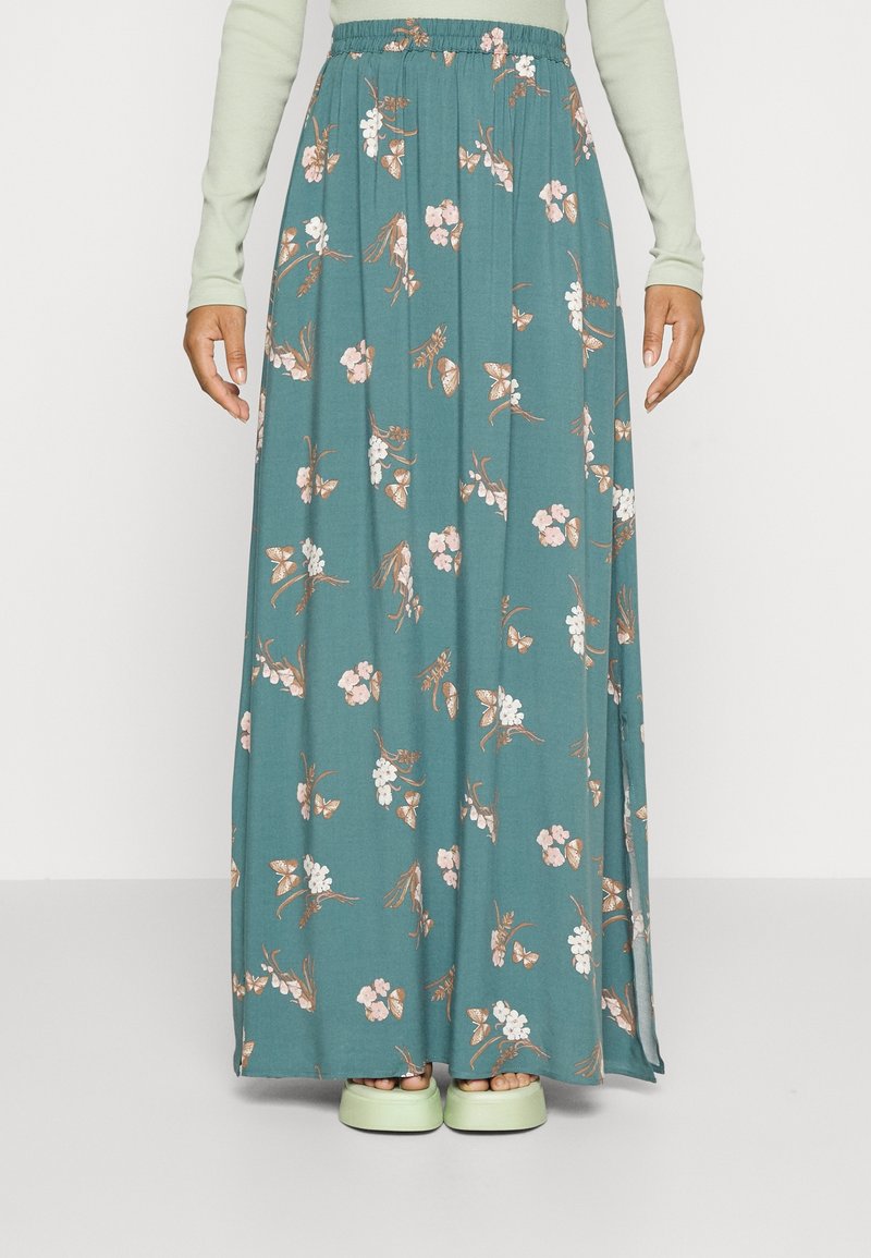 Jupe maxi en tissu teal avec des motifs floraux et de papillons. Dotée d'une taille élastique et d'un design plissé. Portée avec des chaussures vert clair.
