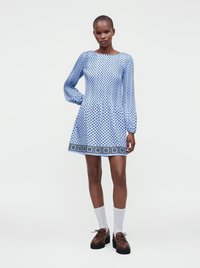 Femme portant une robe bleu clair à manches longues avec des motifs en losange noirs et une bordure décorative, assortie à des chaussettes blanches et des chaussures marron.