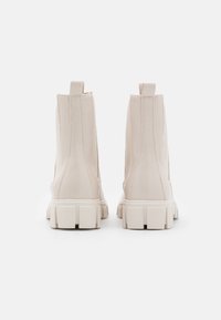 Beżowe skórzane botki za kostkę o zarysowanym fasonie, z widocznymi przeszyciami i grubą, fakturowaną białą gumową podeszwą.