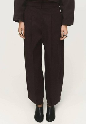 Pantalones de pierna ancha burdeos con pliegues frontales, diseño a medida y textura suave. Combinados con zapatos negros de punta cerrada.