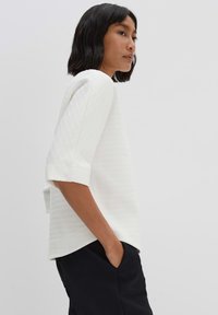 Top bianco con texture e maniche corte, con vestibilità rilassata e tasca laterale, abbinato a pantaloni neri. Design pulito e minimalista.