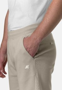 Pantaloni jogger beige con una texture morbida, dotati di tasche laterali inclinati e di un logo ricamato sulla coscia sinistra. Include una cintura elastica.