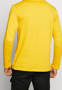 Gelbes Langarmshirt mit glatter Textur, runder Ausschnitt und figurnahen Ärmeln. Einfaches Design ohne Muster oder Akzente.