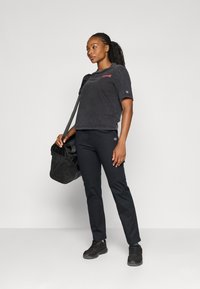 Grå t-shirt med korte ærmer og rødt logo, parret med tætsiddende sorte bukser og sorte sneakers. Bærer en sort duffelbag med netdetaljer.