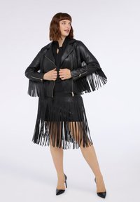 Giubbotto di pelle nera con dettagli in frange sulle maniche e sull'orlo, abbinato a una gonna in frange coordinata. Tacchi neri completano il look.