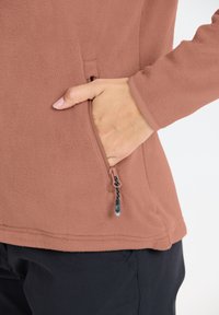 Fleecejacka i dammig rosafärg med detaljer av dragkedjor i fickor. Mjuk textur, rund halsringning och långa ärmar med kontrasterande mudd.