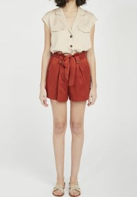 Beige kortärmad skjorta med krage och två bröstfickor; röda högmidjade shorts med rosett och knappdetaljer; platta sandaler.
