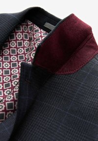 Veste de costume texturée bleu marine avec un col en feutre bordeaux. L'intérieur présente un motif géométrique dans les couleurs bordeaux, gris et crème.