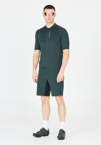 Dunkelgrünes Radoutfit bestehend aus einem kurzärmeligen Trikot mit Reißverschluss vorne, passenden Shorts und schwarzen Radschuhen. Reflexionssonnenbrille.