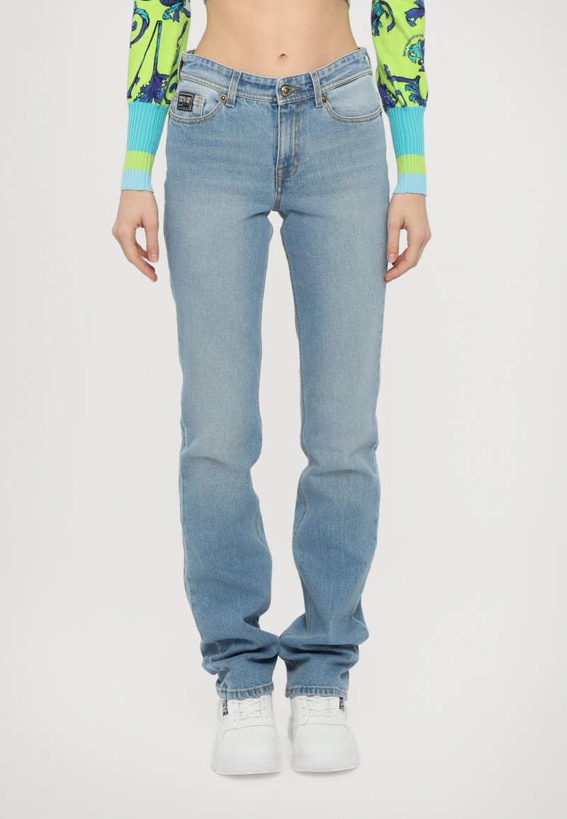 Svetlo modri denim jeans s ravnim krojem, zasnovo s petimi žepi in subtilno teksturo. Nosi se z belimi športnimi čevlji za kontrast.