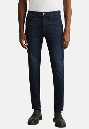 Jeans din denim de culoare albastru închis, cu un croi slim, prezentând cusături portocalii, cinci buzunare și un design curat. Purtați cu pantofi din piele negri.