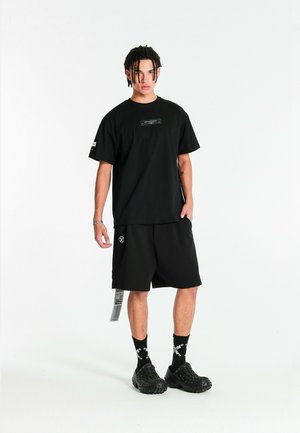 Joven con trenzas que lleva una camiseta negra oversize, pantalones cortos negros con etiqueta, calcetines negros con patrón y zapatos negros gruesos, de pie.