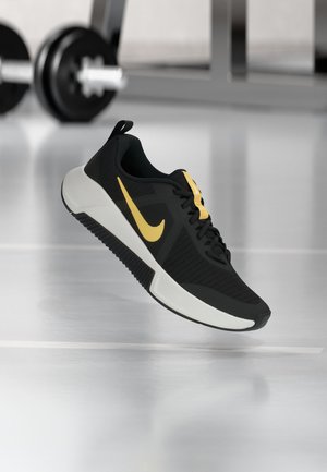 Crno-žuta Nike tenisica lebdi iznad podne površine teretane, s zamagljenom opremom za dizanje utega u pozadini.