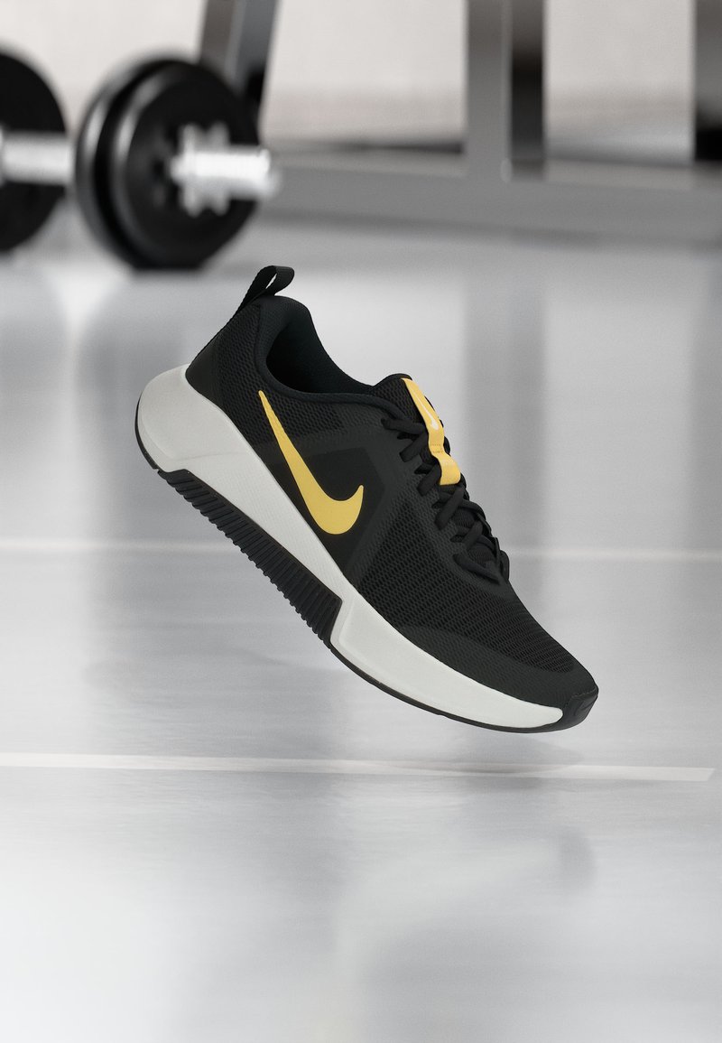 Μαύρο και κίτρινο αθλητικό παπούτσι Nike που αιωρείται πάνω από το δάπεδο γυμναστηρίου, με θολό εξοπλισμό άρσης βαρών στο βάθος.