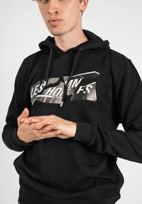 Sudadera negra de algodón con un gráfico frontal que presenta un diseño en capas y desgastado. Tiene un bolsillo tipo canguro en la parte frontal y capucha con cordón.