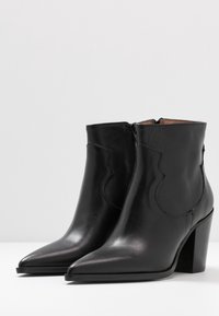 Un par de botas de cuero negras hasta el tobillo con puntas afiladas, tacones cuadrados, cremallera lateral y costuras decorativas sobre una superficie blanca.