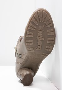 Beige kotníková bota s kulatou špičkou, bočními zipy, texturovaným koženým povrchem, gumovou podrážkou s protiskluzovým vzorem a logem výrobce.