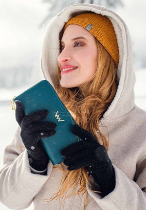 Teal wallet with gold hardware, held in black gloved hands, against a snowy backdrop; person wears a beige jacket and mustard knit beanie.