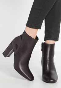 Ciemnobrązowe skórzane botki za kostkę z ostrym noskiem i masywnym, wysokim obcasem. Posiadają boczny zamek błyskawiczny oraz błyszczący akcent na jednej z botków.